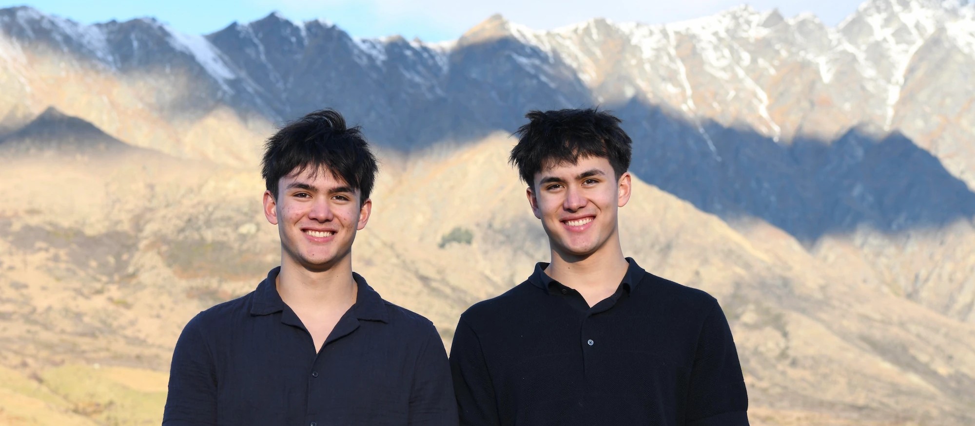 Two brothers standing side by side from the waist up, smiling while gardening with tools in hand.